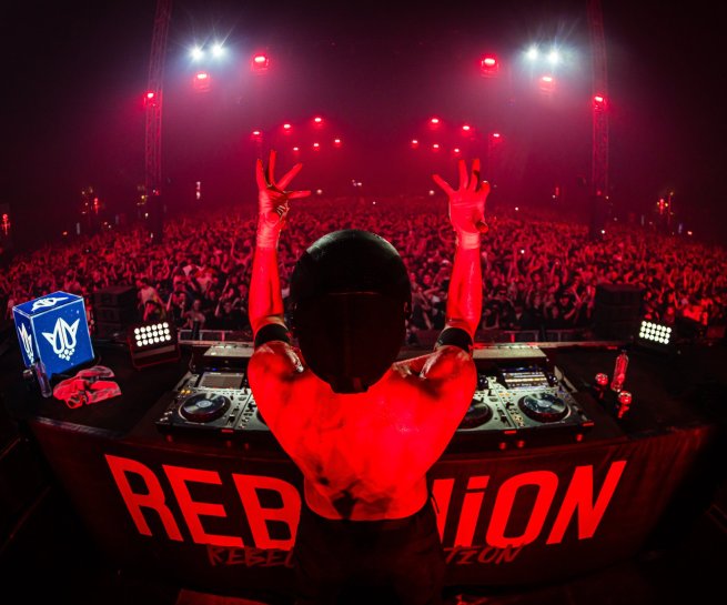 Crowd raises hands while red pyrotechnics burst at REBELLiON Outdoor Festival
